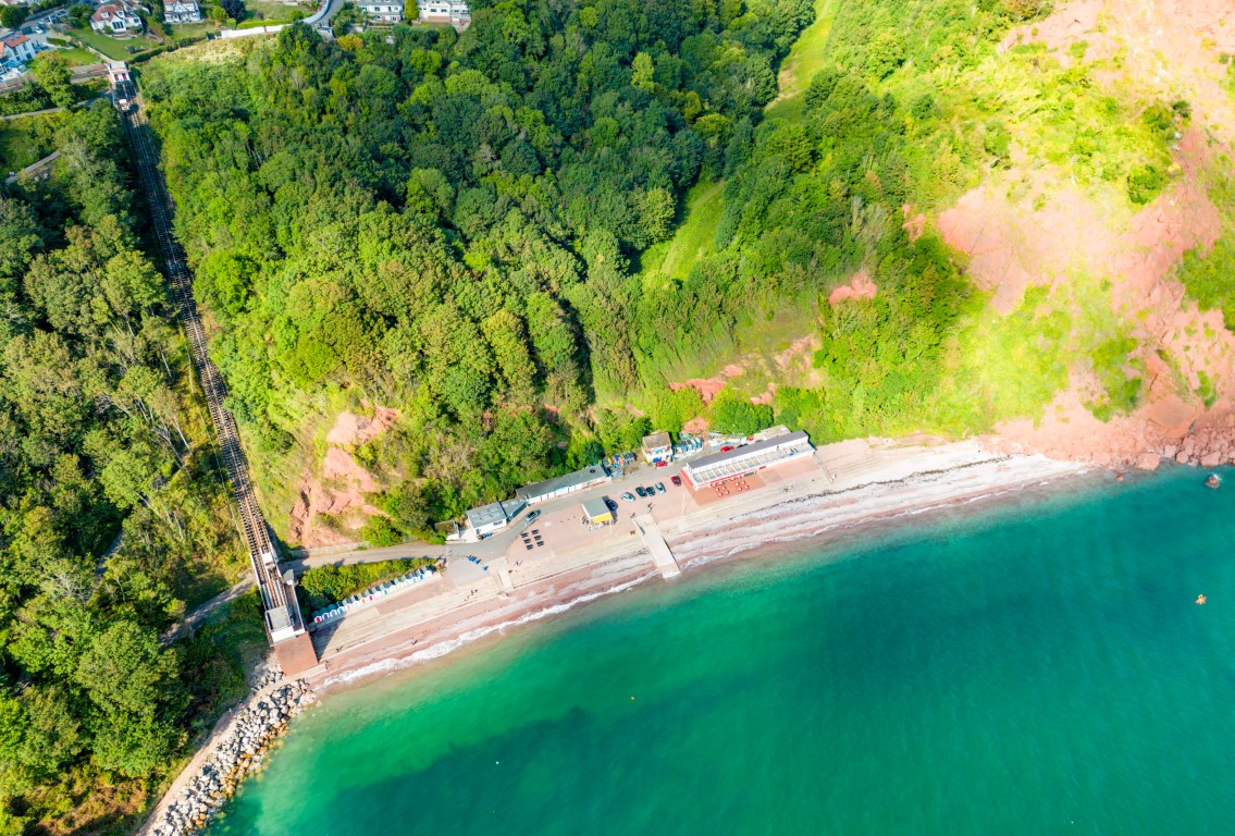 Aerial view of Oddicombe Beach, with the cliff railway, beach huts and turquoise water beneath the red cliffs